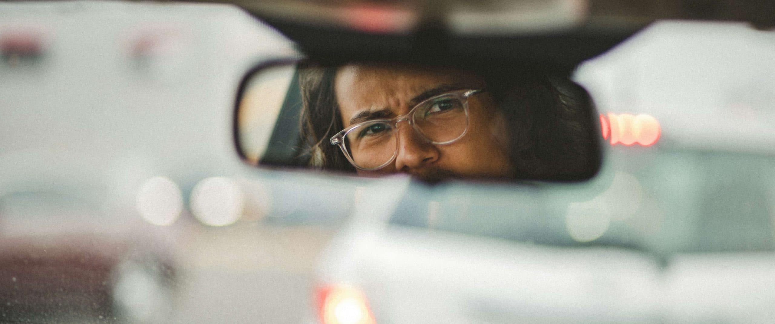 A man in clear-framed glasses, seen in a rearview mirror, in front of a blurred windscreen.