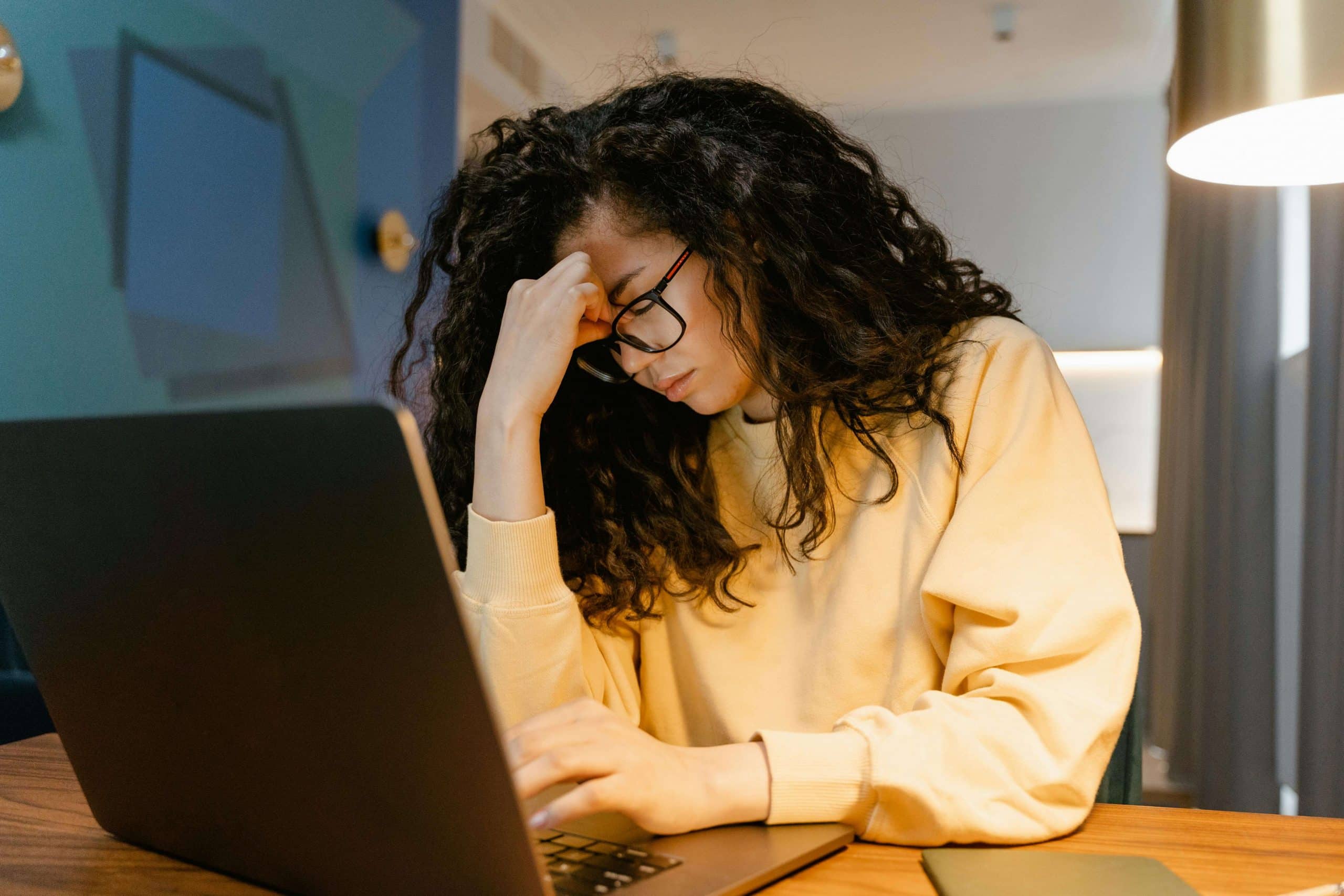 A young woman in glasses sitting at a computer massages her tired eyes, representing the need for anti-fatigue lenses.
