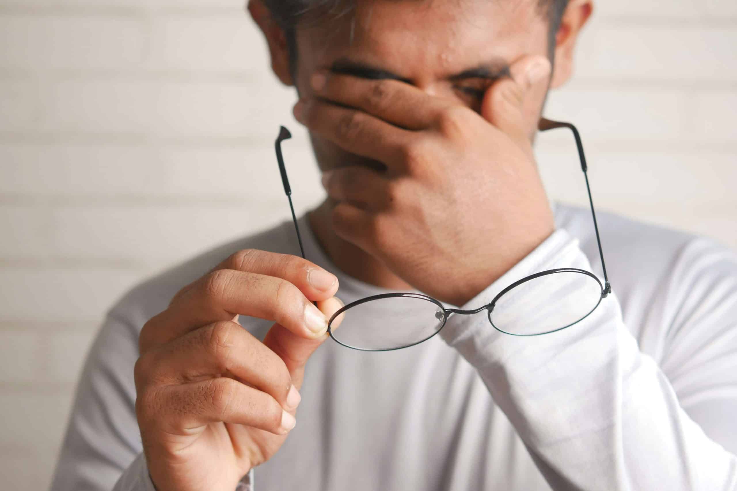 A man in a white shirt rubbing his tired eyes while holding his glasses, illustrating the discomfort people can feel when trying to get used to varifocals.